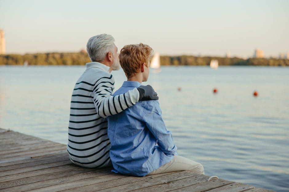 Elderly man and young boy seated on a dock, embracing while observing a serene lake.